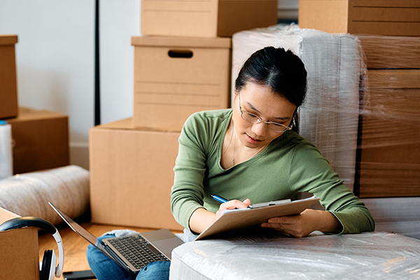 A woman sits in her living room, using a laptop and a clipboard to keep track of her long-distance moving tasks. 